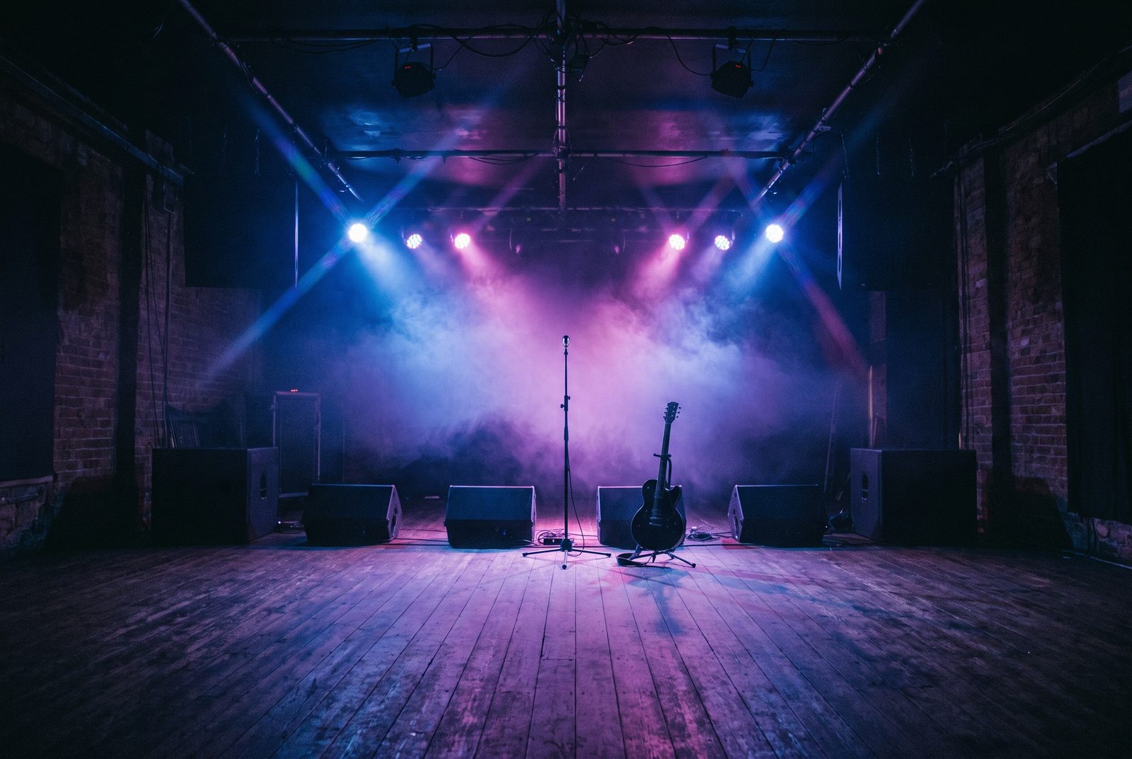 An empty club stage lit in blue and magenta, with a microphone stand and electric guitar waiting